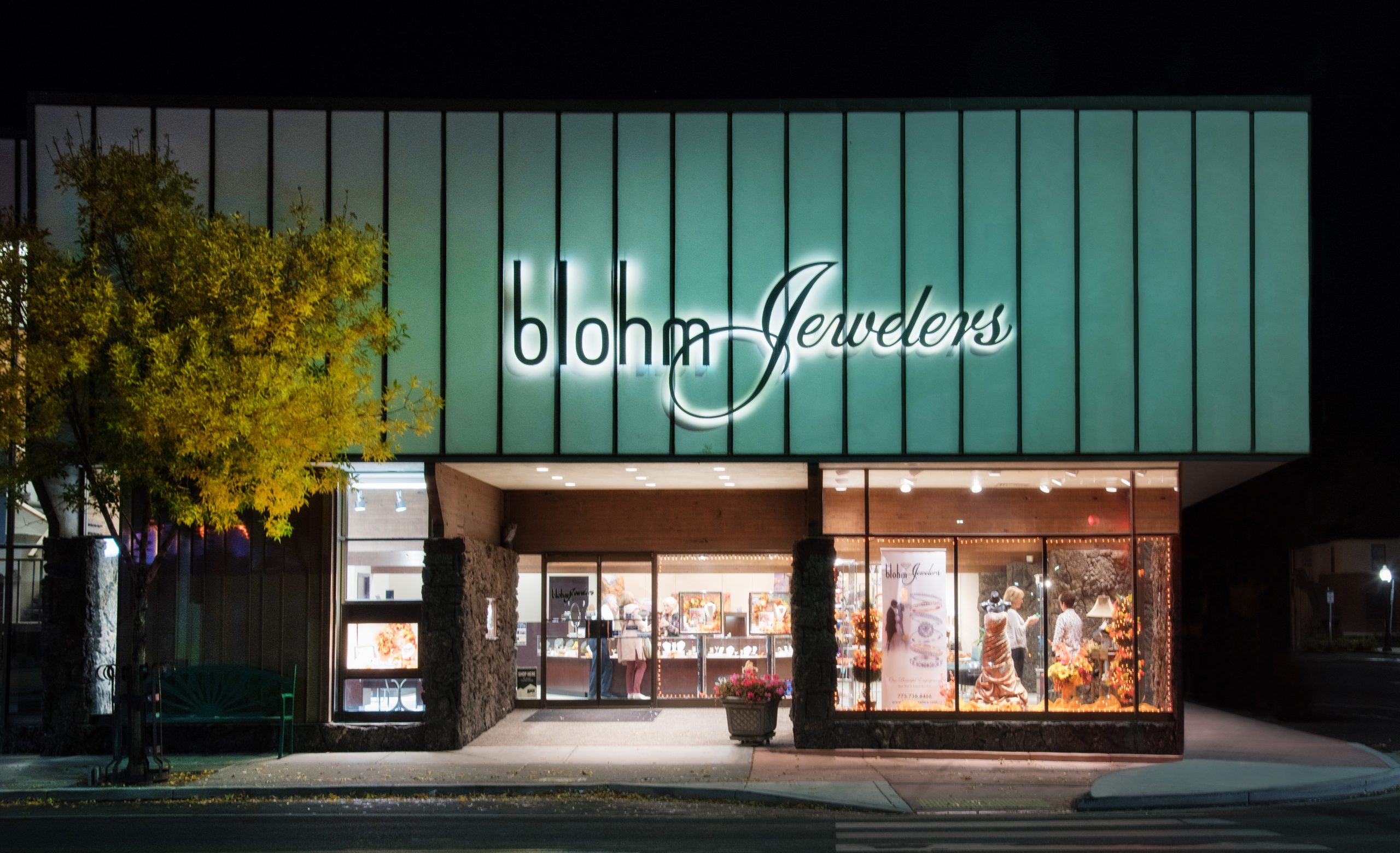 Blohm Jewelers storefront at night, featuring illuminated signage and fall decorations. Displays showcase jewelry