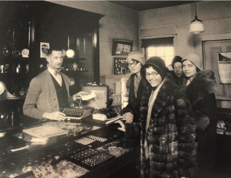 A man behind a store counter interacts with four fashionable women in fur coats